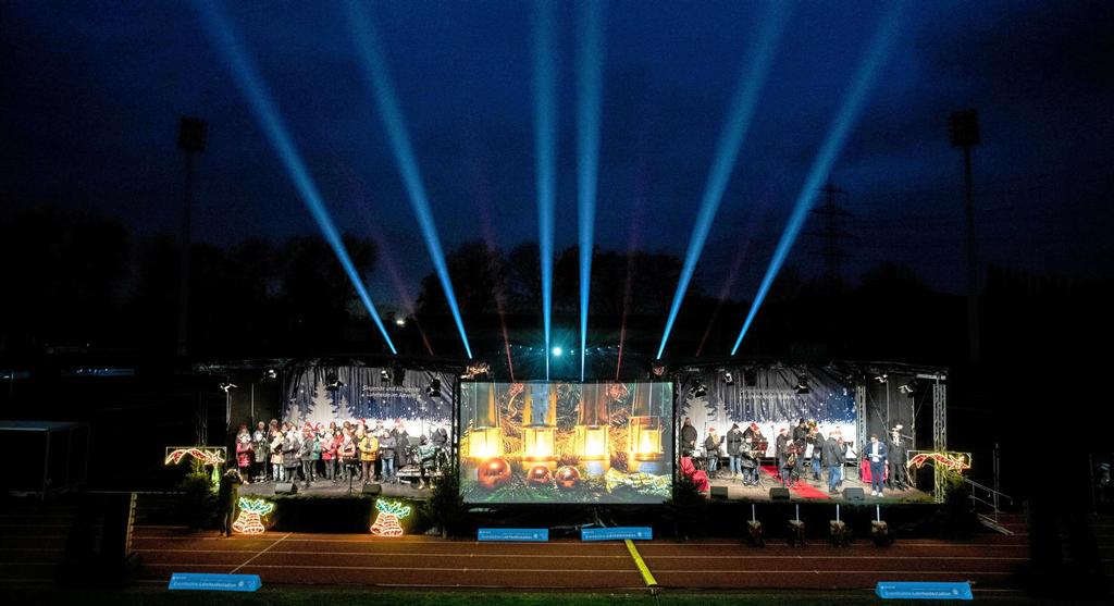 Foto: Zwei beleuchtete Bühnen, in der Mitte eine Leinwand, im Hintergrund 6 Strahler in den Nachthimmel.